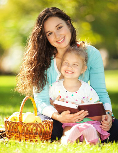 a smiling mother and young daughter enjoy reading a book outside on a summer day